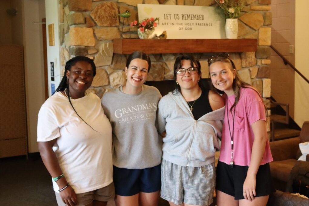 Four smiling young women standing in front of a sign that reads "Let us remember we are in the holy presence of God."