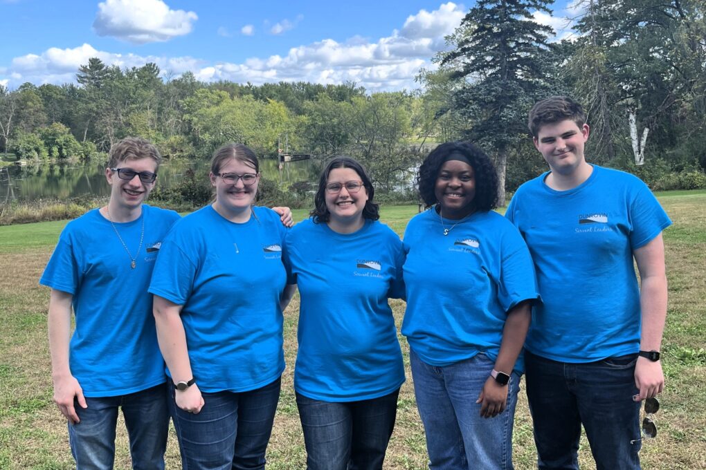 Five young adults in bright blue shirts standing together with a blue sky, green trees, and pond in the background