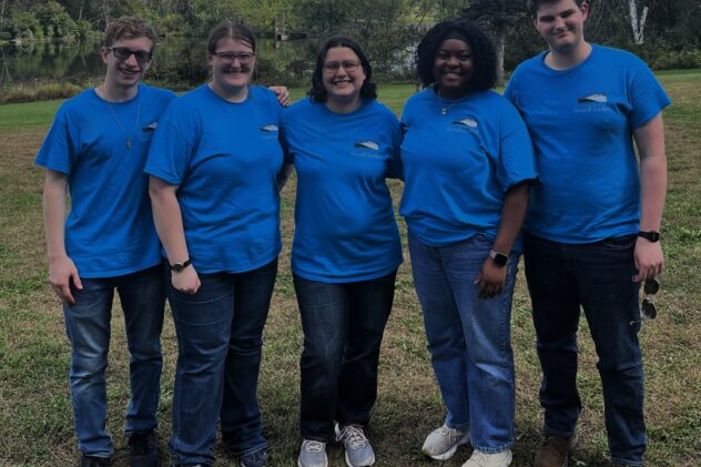 Five young adults wearing blue shirts with green grass and trees in the background