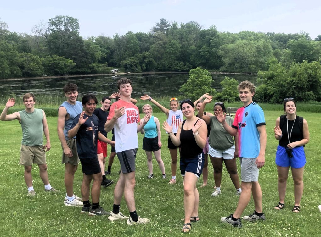 A group of racially diverse high school students on a green lawn with water and green trees in the background