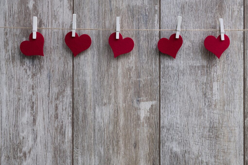 A grey barnwood wall with five small, red Valentine hearts clipped to twine by clothespins